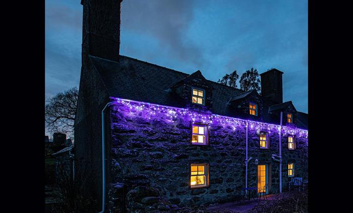 Betws Bach Farmhouse with Festive Lights hanging along the front. Blue icicles on at evening with a dark sky and lights on, inside the farmhouse